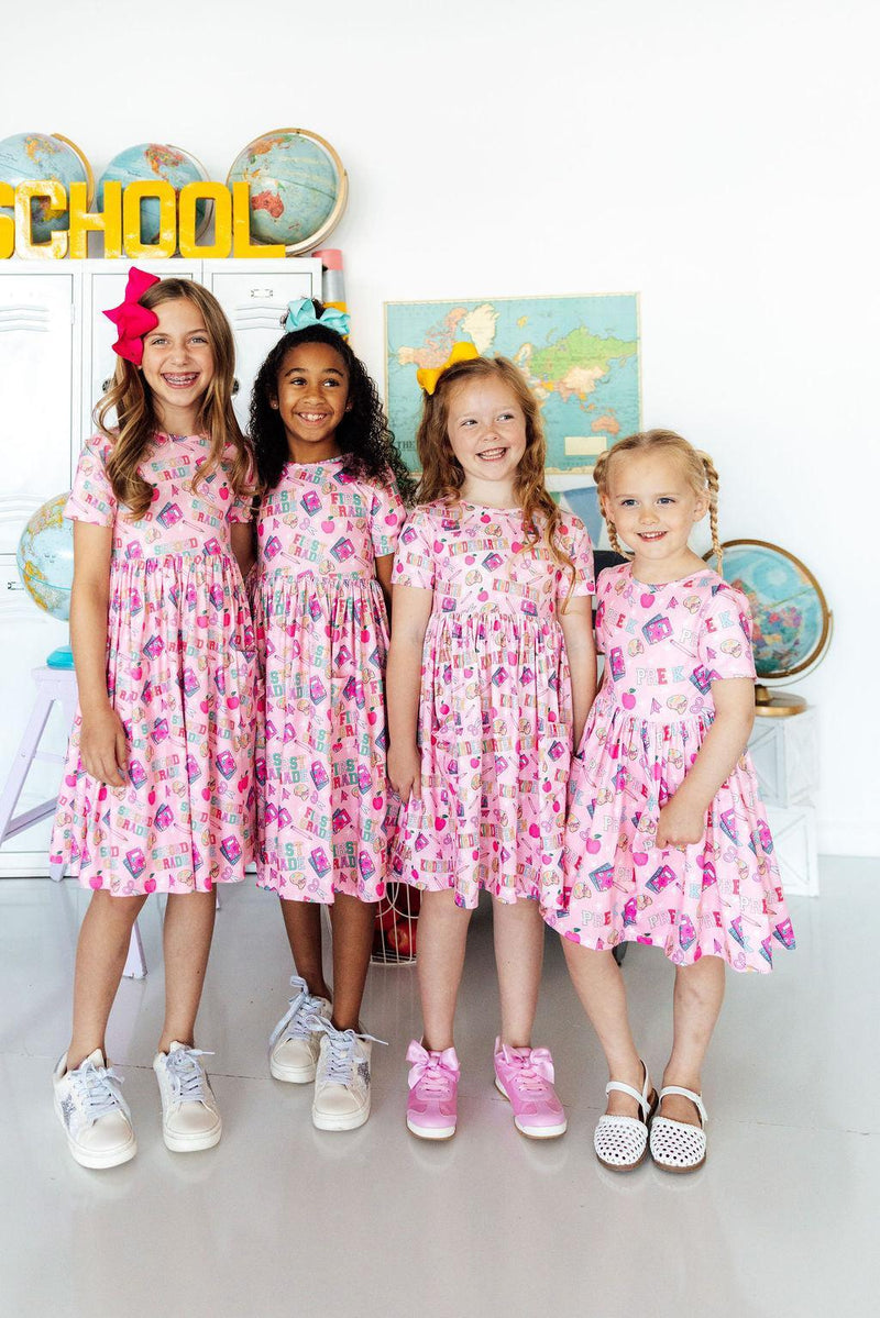Four girls in pink 2nd-grade short-sleeve pocket twirl dresses, twirling in a classroom.