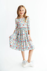 Child wearing a multi-color floral 3/4 sleeve twirl dress with front pockets and white sneakers.