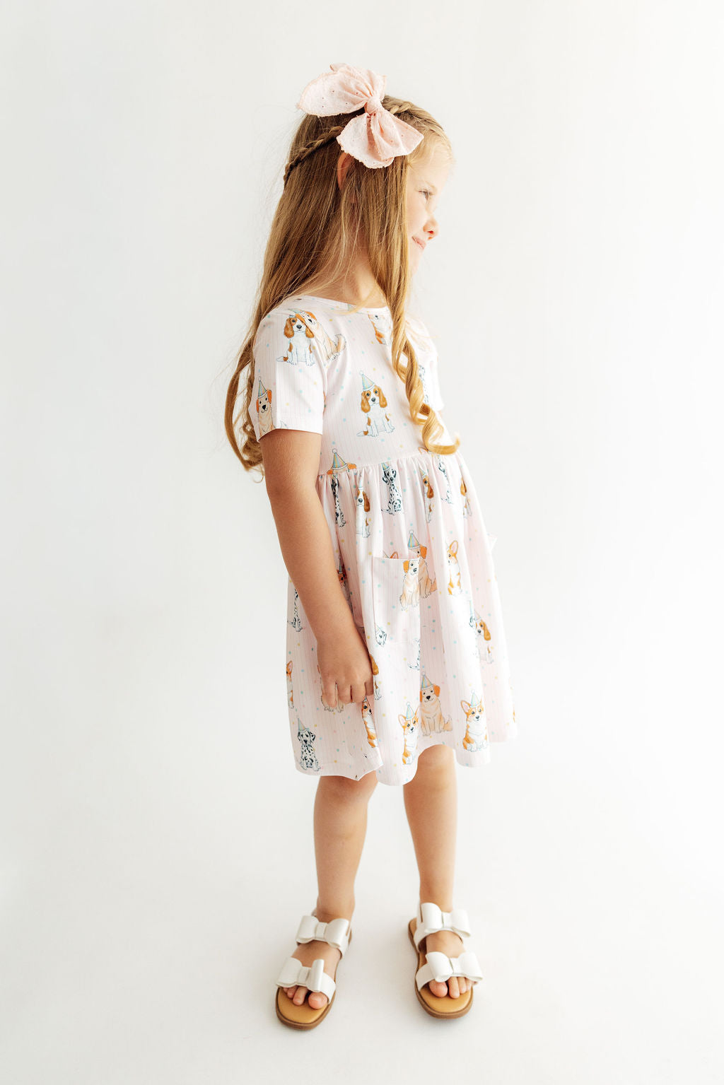 Girl wearing a pink short sleeve twirl dress with puppy print, front pocket, and matching bow. Designed for comfort and easy play.