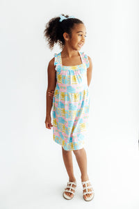 Girl wearing Tropical Summer Ruffle Cross Back Dress with pastel print and ruffled shoulder straps.