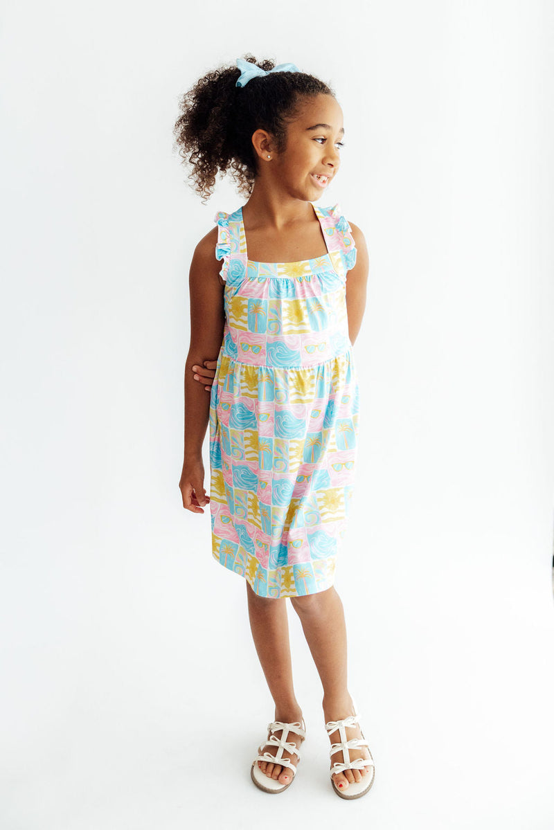 Girl wearing Tropical Summer Ruffle Cross Back Dress with pastel print and ruffled shoulder straps.