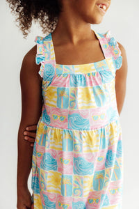 Girl wearing Tropical Summer Ruffle Cross Back Dress with pastel palm-sun print and ruffle straps.