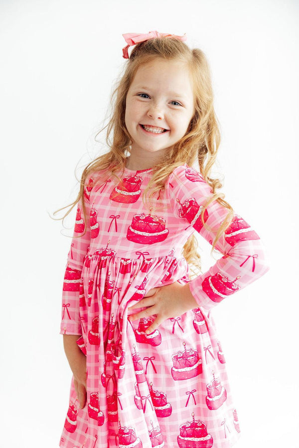 Smiling little girl in Make a Wish L/S Pocket Twirl Dress, pink cake-print long-sleeve twirl dress with a bow.