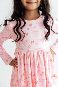 Pink long sleeve twirl dress with dreidel, menorah, and Star of David print, featuring side pockets and a full circle skirt.