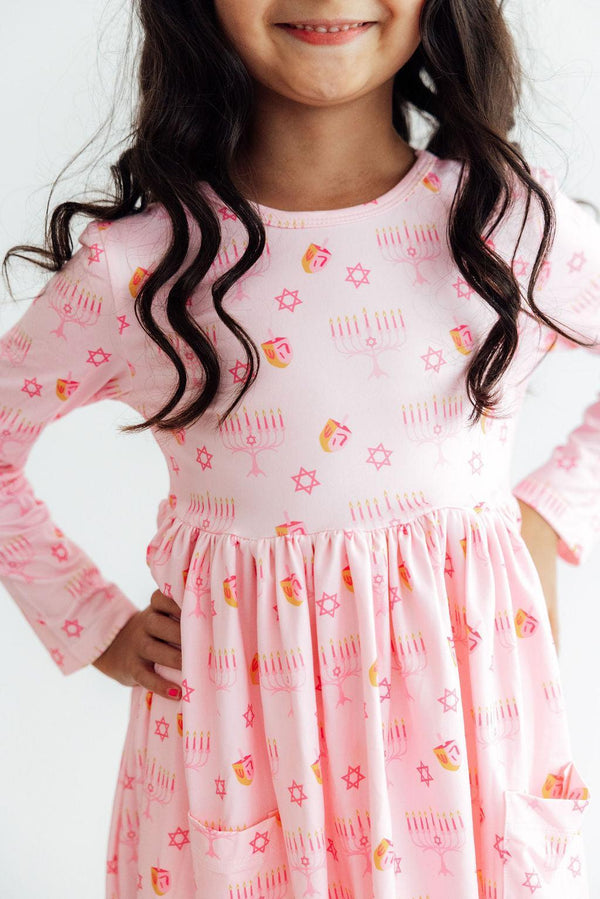 Pink long sleeve twirl dress with dreidel, menorah, and Star of David print, featuring side pockets and a full circle skirt.