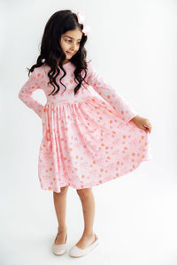Child twirling in a pink long sleeve dress featuring dreidel and menorah prints, full circle skirt, front pockets, and a matching hair bow.