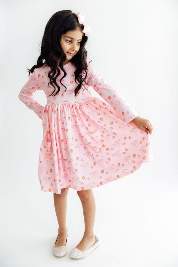 Child twirling in a pink long sleeve dress featuring dreidel and menorah prints, full circle skirt, front pockets, and a matching hair bow.