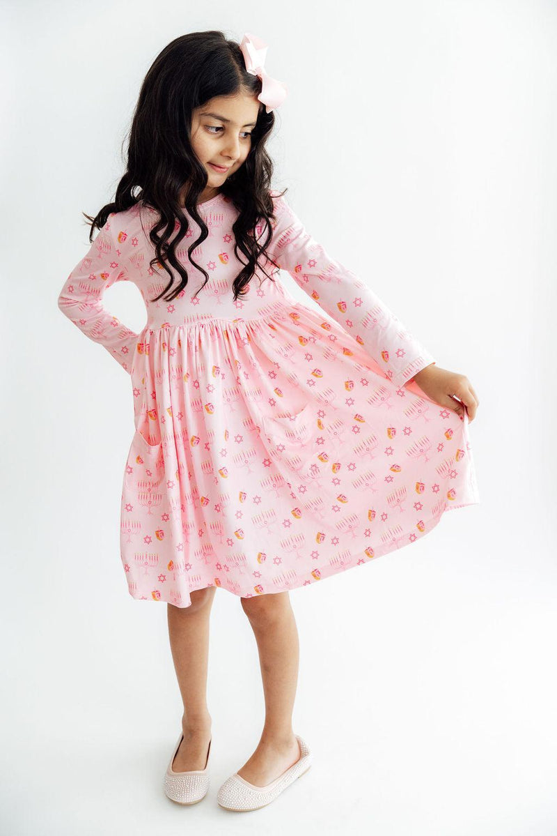 Child twirling in a pink long sleeve dress featuring dreidel and menorah prints, full circle skirt, front pockets, and a matching hair bow.