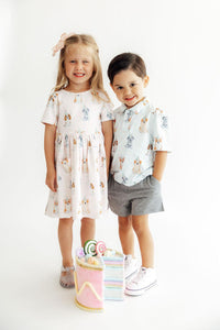Girl in pink dog print twirl dress and boy in matching puppy print shirt stand with felt cake. Comfortable, easy-wash party pups childrenswear.