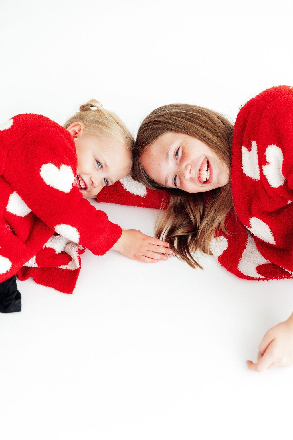Mother and daughter lying together laughing in matching red and white heart cozy cardigans.