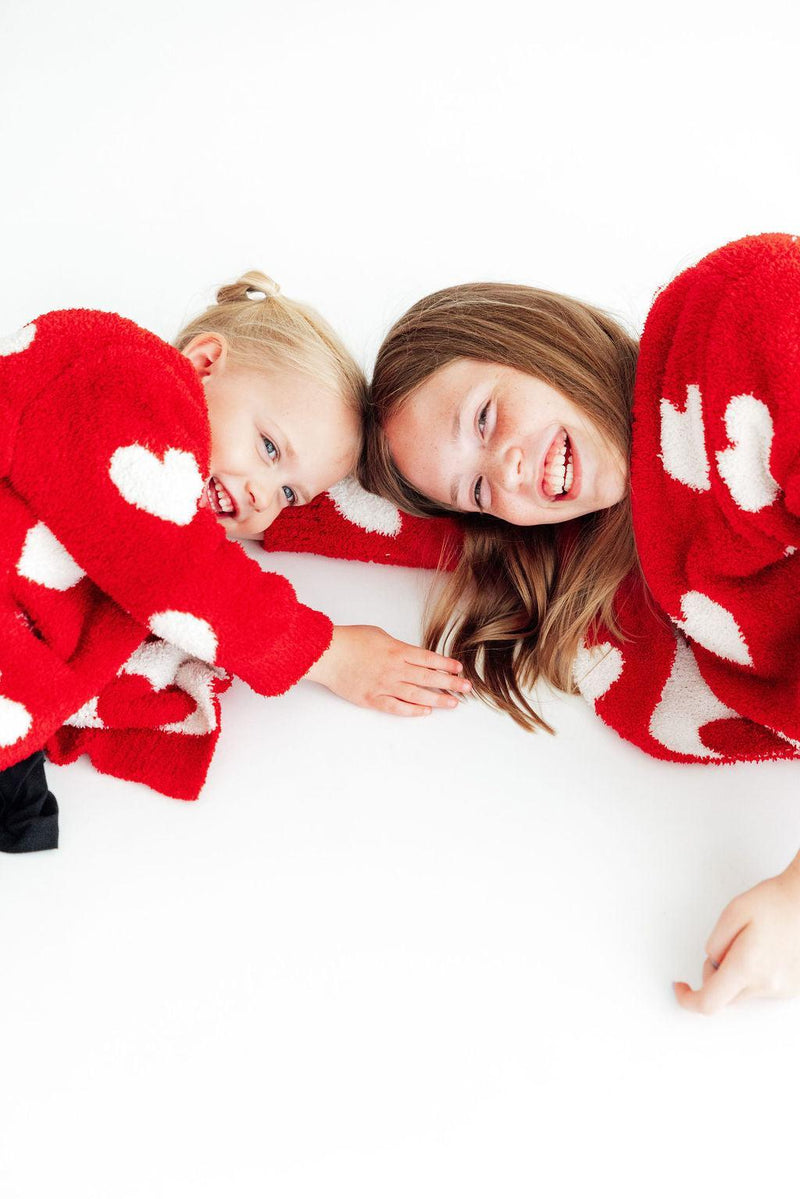 Mother and daughter lying together laughing in matching red and white heart cozy cardigans.