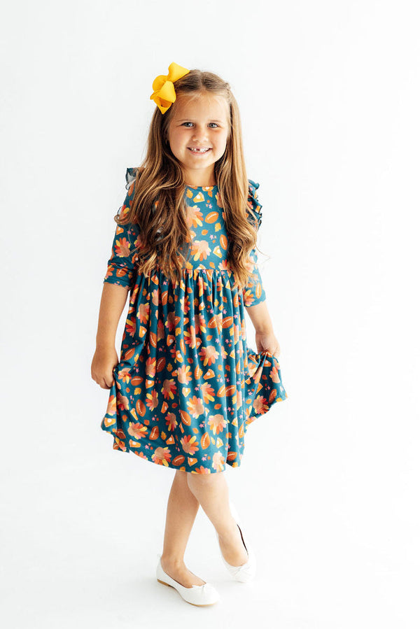 Gobble til You Wobble 3/4 Sleeve Ruffle Twirl Dress in teal floral, on a smiling girl with a yellow bow.