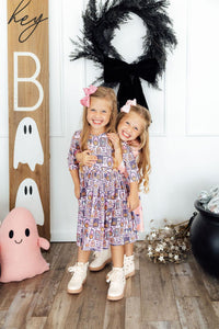 Two smiling girls pose in Halloween print dresses near ghost decor. Features 3/4 sleeves, pockets, and a twirl skirt designed for comfort.