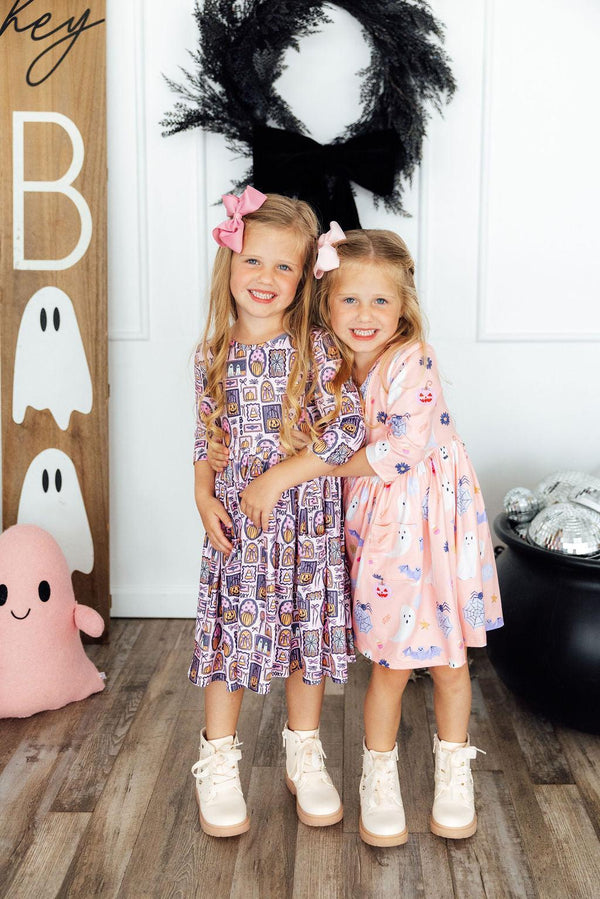 Two girls hug wearing Halloween print 3/4 sleeve pocket twirl dresses in purple haunted house and pink ghost patterns with white boots.