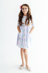 Girl wearing pastel mermaid scale print 3/4 sleeve pocket twirl dress with full skirt, scoop back, and ballet neckline.