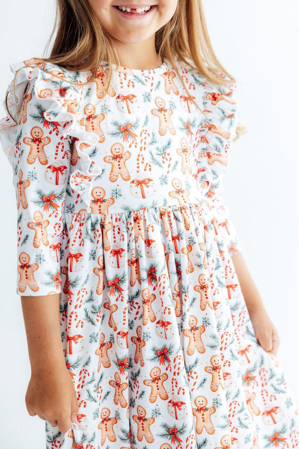 Girl wearing white 3/4 sleeve ruffle twirl dress with gingerbread men, candy canes, and pine print for Christmas.