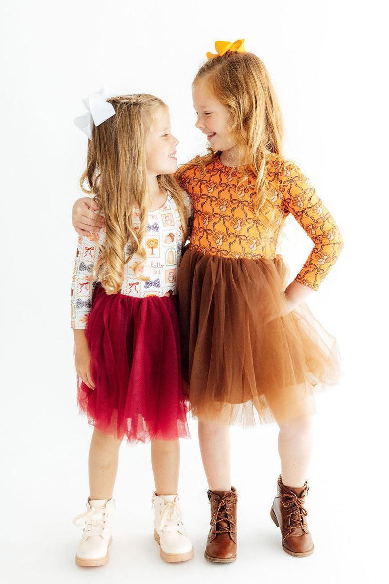 Two girls modeling long sleeve tutu dresses in autumn colors. One features a fall print top with a burgundy tulle skirt, the other has an orange lace bodice with a brown tulle skirt.