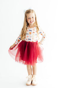 Little girl wearing Hello Fall long sleeve tutu dress with autumn print top and red layered skirt, paired with cream boots and white bow.