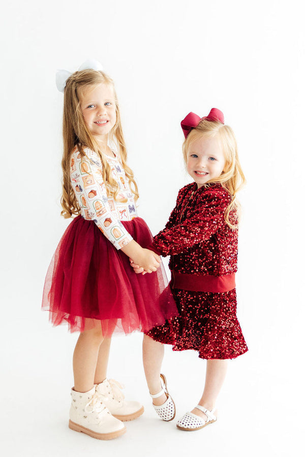 Two girls holding hands wearing fall outfits: one in a long sleeve printed top with a red tulle tutu dress and white boots, the other in a red sequin set with a bow.