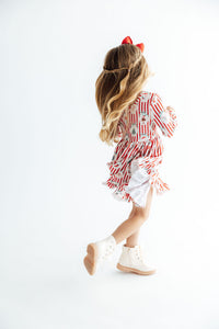 Back view of girl twirling in red striped Santa Sweet Shoppe dress with ruffle hem, white boots, and red bow.