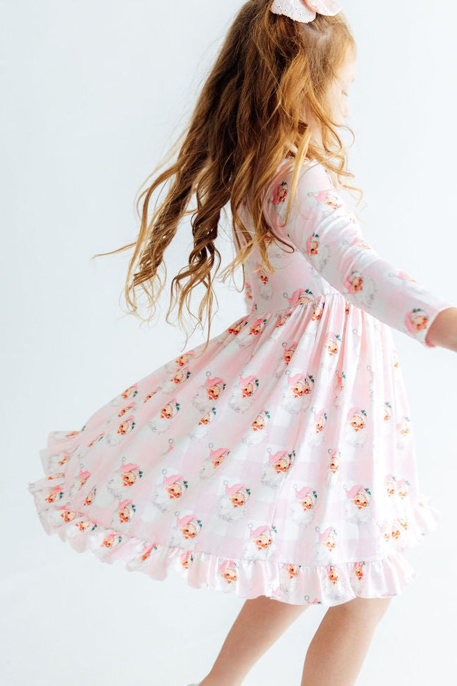 Girl twirling in a pink gingham long sleeve ruffle hem dress printed with Storybook Santa motifs.