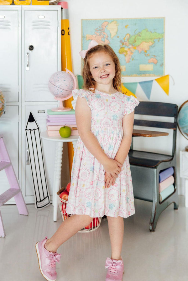 Girl wearing Apple of My Eye Flutter Sleeve Twirl Dress in pastel print, pink sneakers, classroom backdrop.
