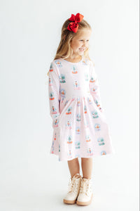 Girl in pink long sleeve pocket twirl dress with snow globe print, red bow hair accessory, and white boots.
