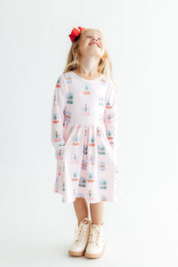 Girl twirling in pink long sleeve pocket dress with snow globe print, red bow hair accessory, and cream boots.
