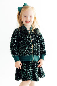 Smiling toddler girl wearing a dazzling hunter green velvet sequin jacket and matching skirt with a gold zipper and green bow.