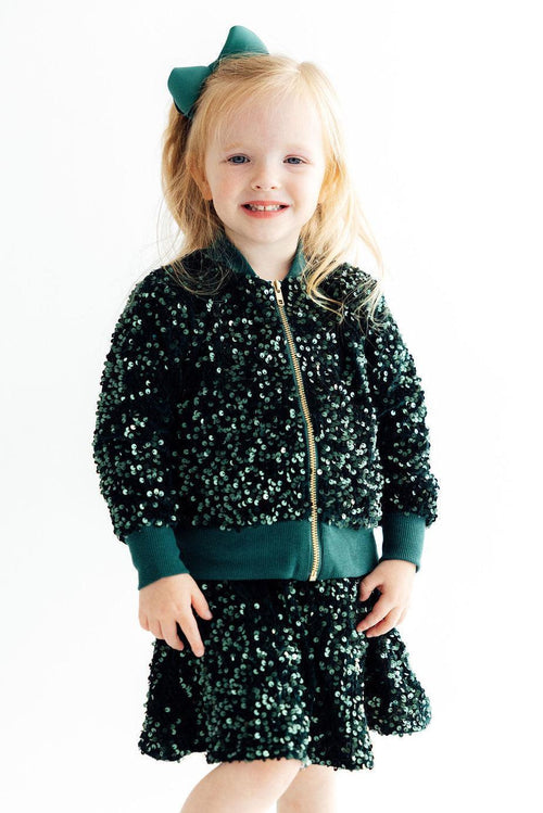 Smiling toddler girl wearing a dazzling hunter green velvet sequin jacket and matching skirt with a gold zipper and green bow.