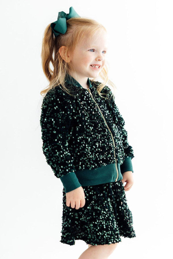 Girl wearing a hunter green velvet sequin jacket and matching skirt with a green bow in her ponytail.