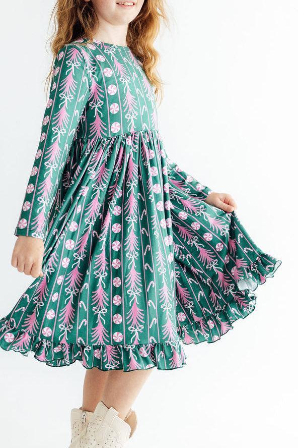 Girl twirling in a green long sleeve ruffle hem dress featuring pink peppermints, candy canes, and holiday trees.