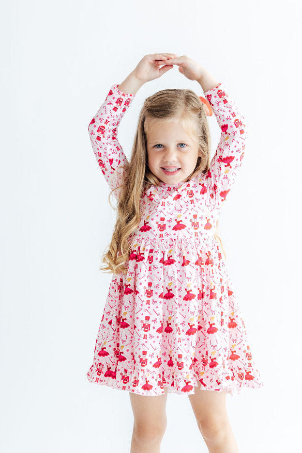 Girl twirling in a long sleeve pink dress with red ballet and nutcracker print, ruffle hem, and full skirt for play.
