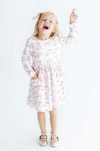 Young girl pointing up while wearing a pink long sleeve dress with unicorn and rainbow print, full skirt, and side pocket.