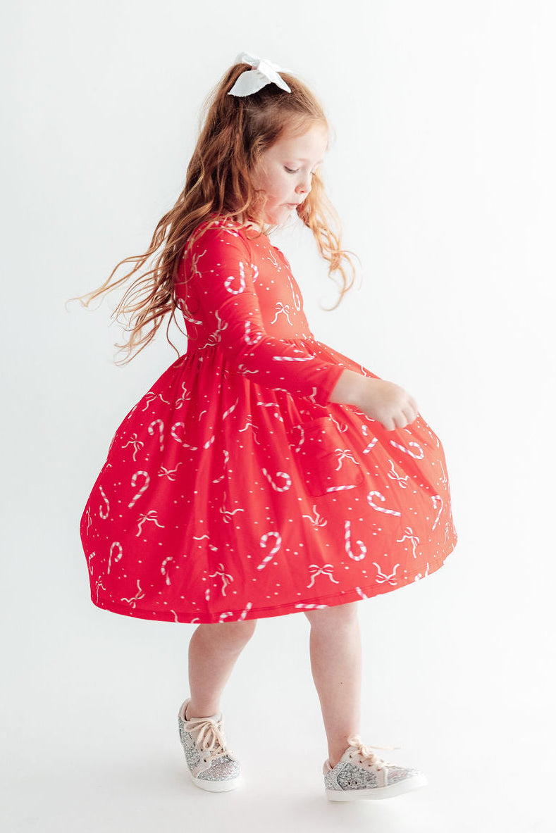 Girl twirling in red long sleeve pocket dress with candy cane print and ballet neckline for festive play.