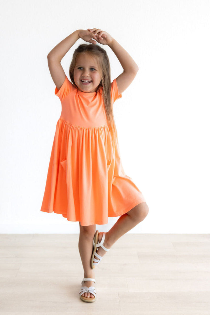 Happy girl dancing in the Neon Coral Short Sleeve Pocket Twirl Dress, demonstrating its twirling capability and bright color.
