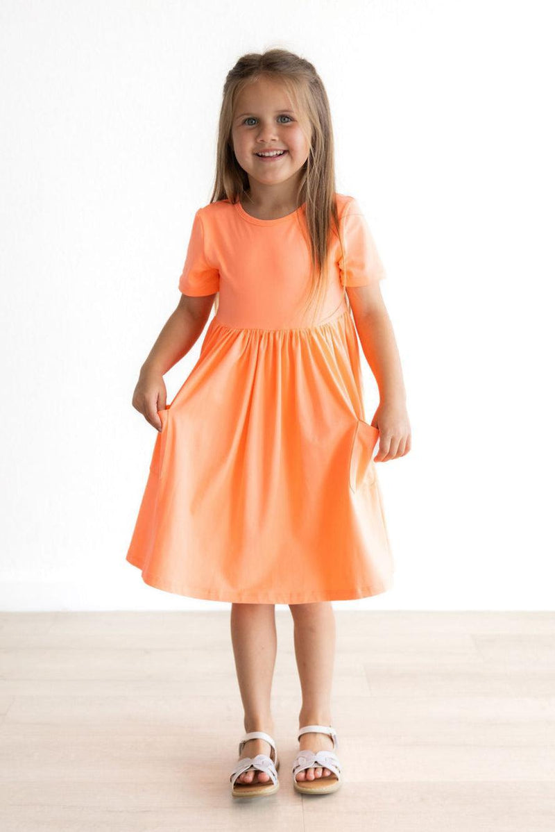 Joyful girl posing in the Neon Coral Short Sleeve Pocket Twirl Dress, with a playful stance showcasing its fun design and practical pockets.