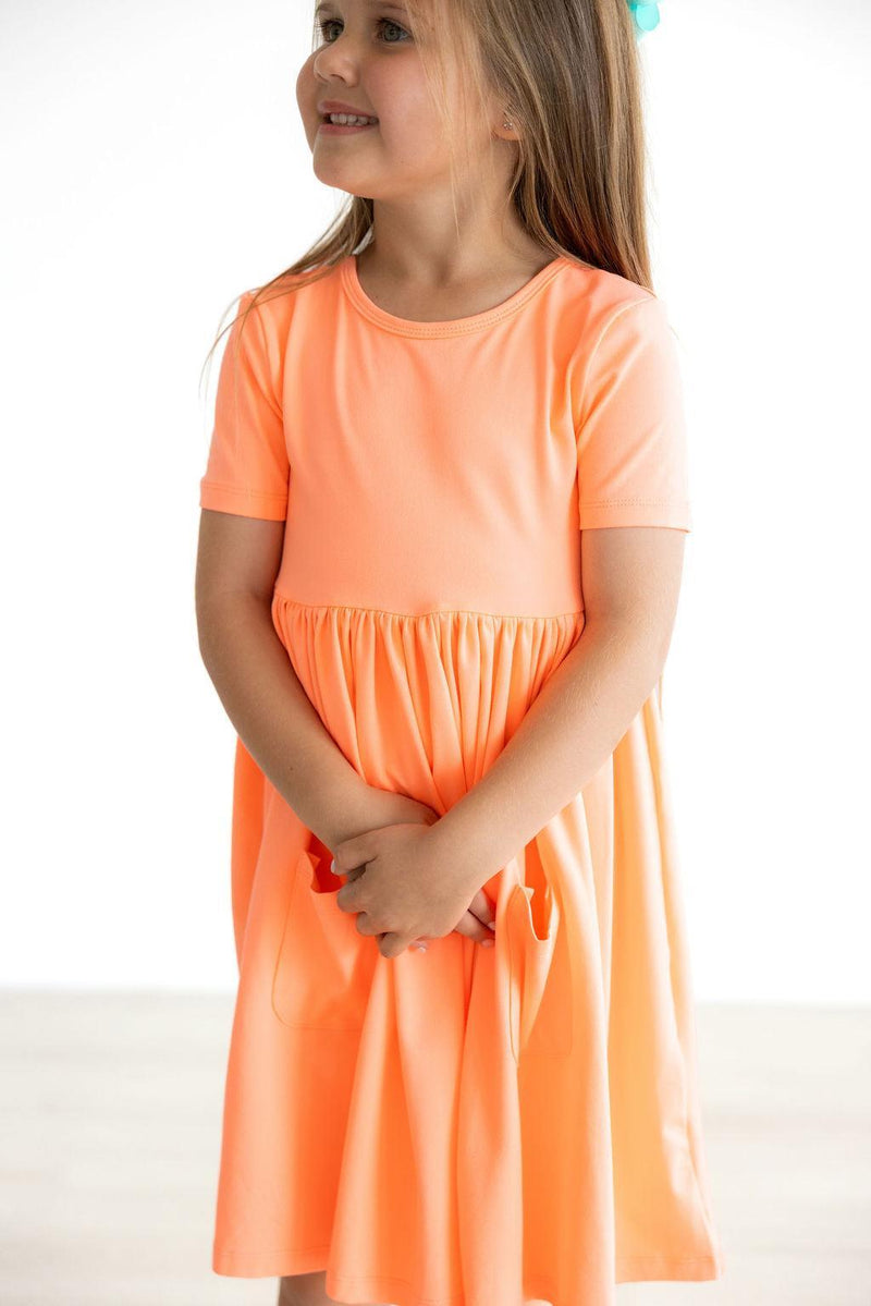 Charming girl in the Neon Coral Short Sleeve Pocket Twirl Dress, holding the dress's pockets and smiling, highlighting comfort and style.