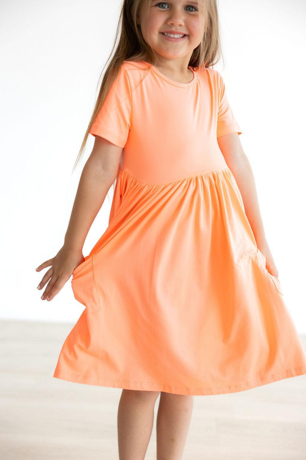 Smiling girl wearing the Neon Coral Short Sleeve Pocket Twirl Dress, showcasing its vibrant color and practical side pockets.