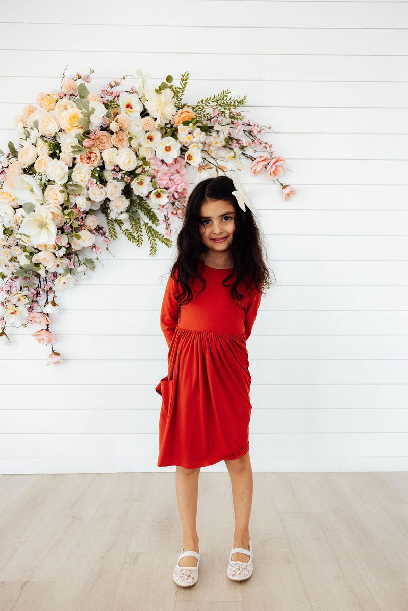 Smiling girl in the Pumpkin Spice 3/4 Sleeve Pocket Twirl Dress, standing against a floral backdrop, radiating autumn charm.
