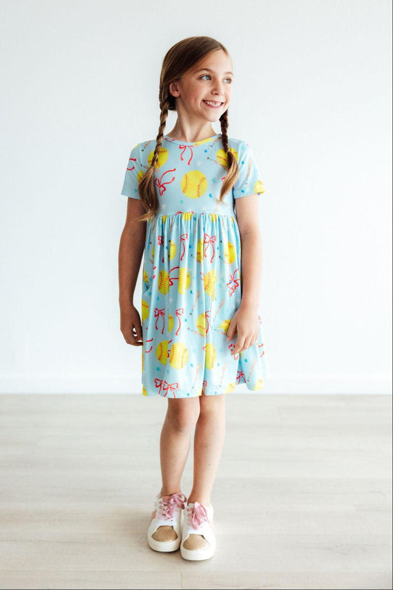 A girl in the Softball Sweetie S/S Pocket Twirl Dress stands confidently, highlighting its playful design with softballs and ribbons, ideal for game days and casual outings.
