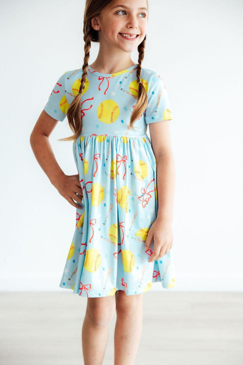 Girl smiling while wearing the Softball Sweetie S/S Pocket Twirl Dress by Mila & Rose, featuring a playful yellow softball and red bow print, perfect for little sports enthusiasts.
