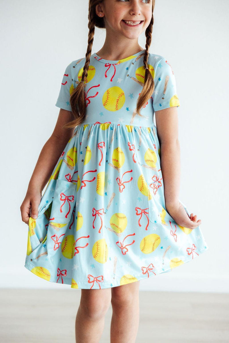 The Softball Sweetie S/S Pocket Twirl Dress being twirled by a happy girl, showing off the fun movement and vibrant softball designs, a must-have for young athletes.