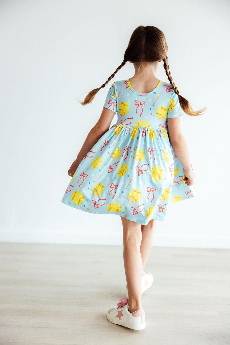 Girl twirling in the Softball Sweetie S/S Pocket Twirl Dress by Mila & Rose, showcasing the cute design with yellow softballs and red bows, ideal for fun and active play.
