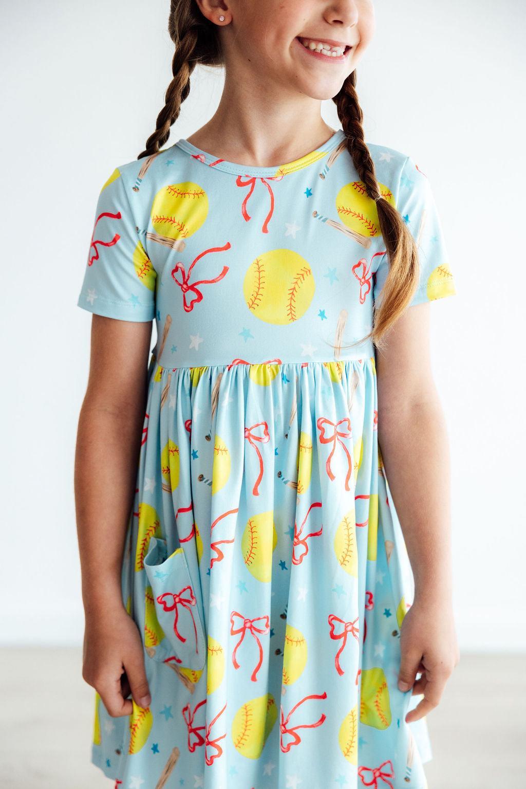 Softball Sweetie S/S Pocket Twirl Dress showcased on a smiling girl, featuring colorful softball and ribbon patterns, perfect for little sports enthusiasts.