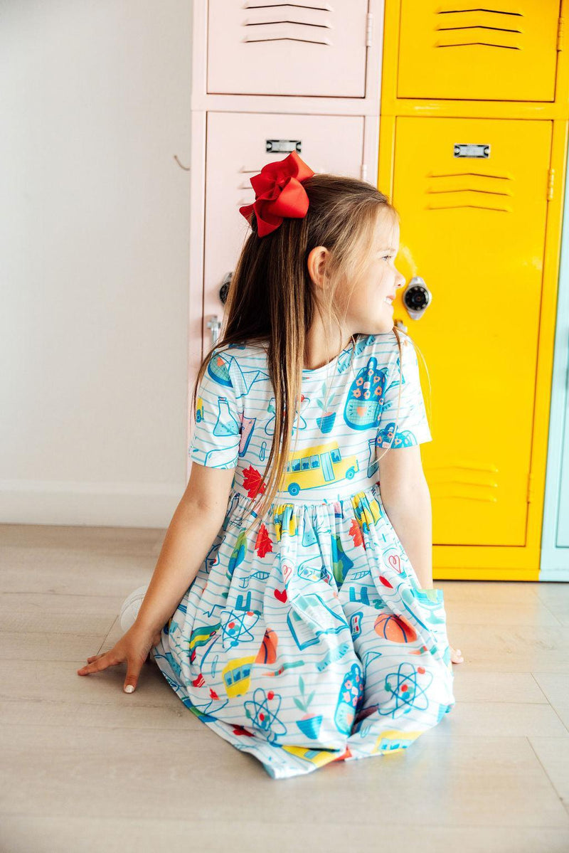 A girl in the School Rules Short Sleeve Pocket Twirl Dress for Girls sits beside lockers, with a charming smile. This twirl dress for girls is designed for comfort and style during school activities.
