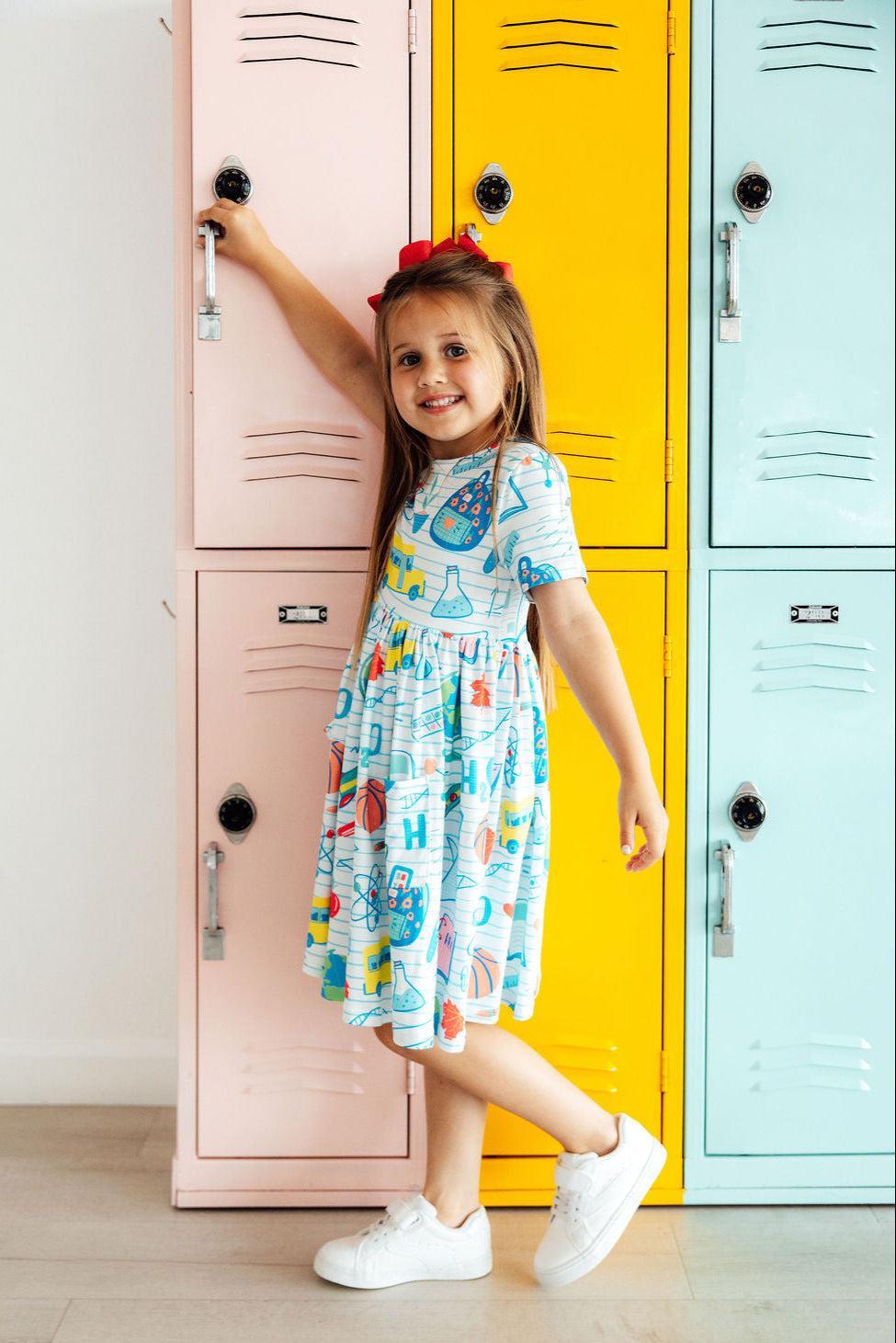 A girl in the School Rules Short Sleeve Pocket Twirl Dress for Girls standing next to colorful lockers, showcasing a fun and playful style, perfect for school and everyday adventures. This twirl dress for girls adds charm to any occasion.