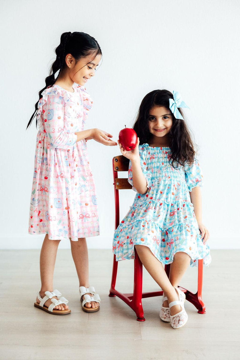 Two girls in the School Days 3/4 Ruffle Twirl Dresses by Mila & Rose ®, one holding an apple and the other playfully reaching out, sitting in a vibrant classroom.