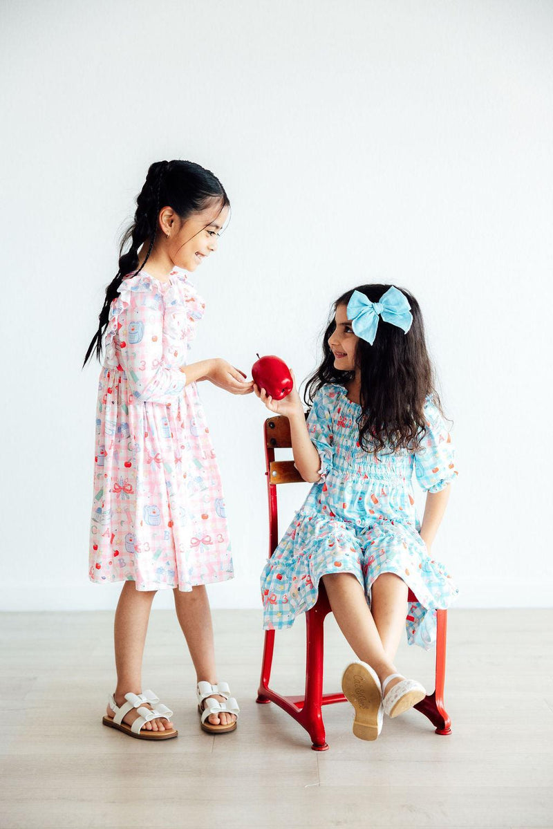 Two friends in charming Honor Roll Smocked Ruffle Dresses by Mila & Rose®, sharing a sweet moment with a bright red apple, highlighting playful fashion.