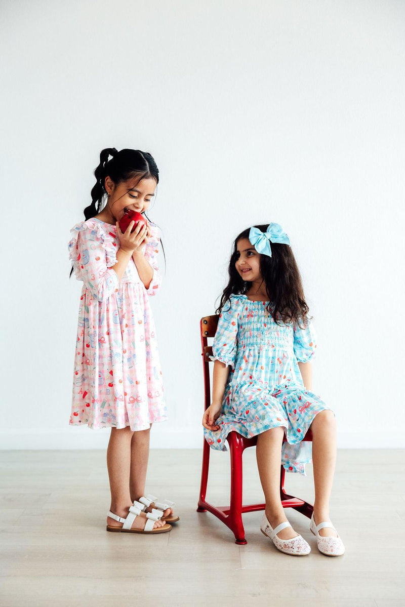Two girls in the School Days 3/4 Ruffle Twirl Dress by Mila & Rose, one sitting on a tiny red chair and the other playfully holding an apple, reflecting the fun and versatile nature of the dress.